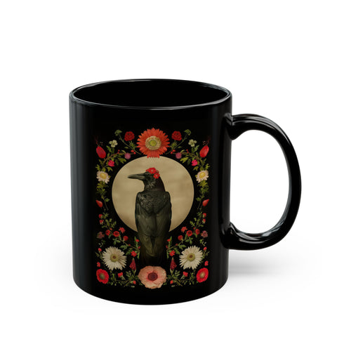 A black ceramic mug featuring a gothic design of a raven against a golden full moon, framed by a floral wreath of coral-colored daisies, white chrysanthemums, and delicate botanical elements. The design includes rich green foliage and small red buds arranged in a symmetrical pattern. The mug has a classic C-shaped handle with a glossy finish.